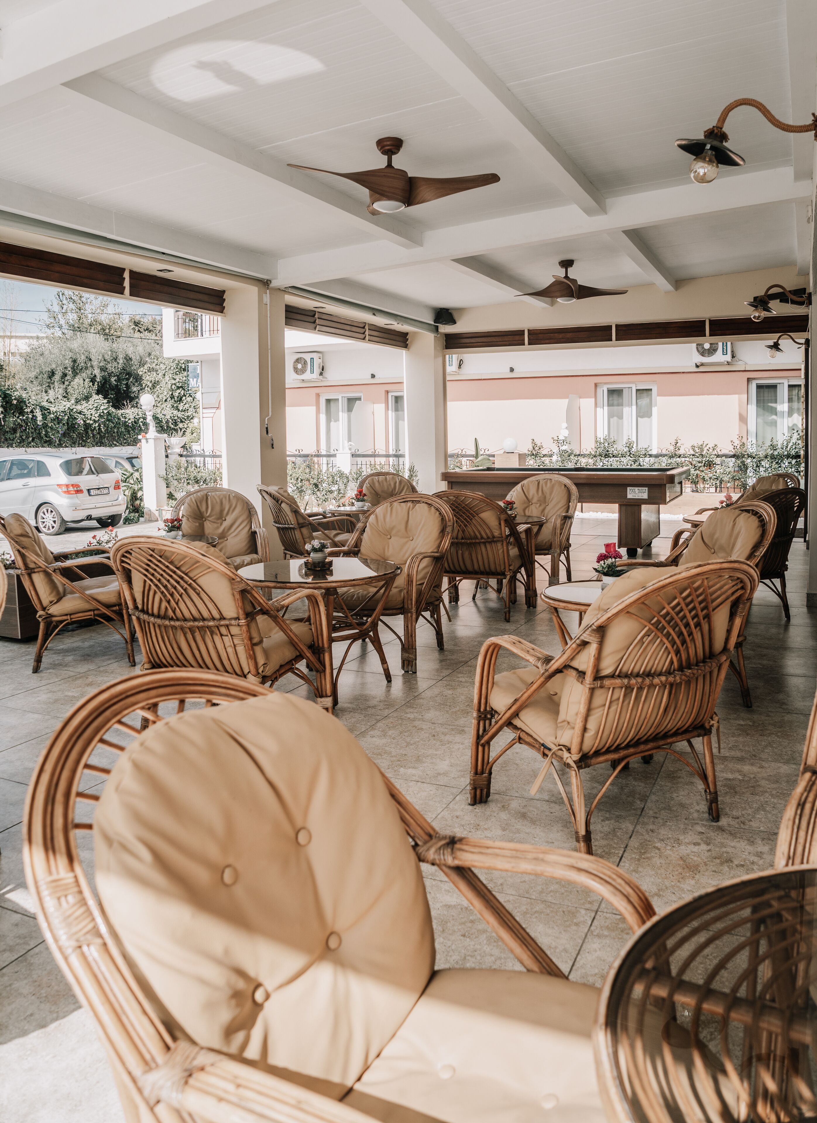 A covered outdoor seating area with wicker chairs and tables, a ceiling fan, and a view of a street with parked cars.