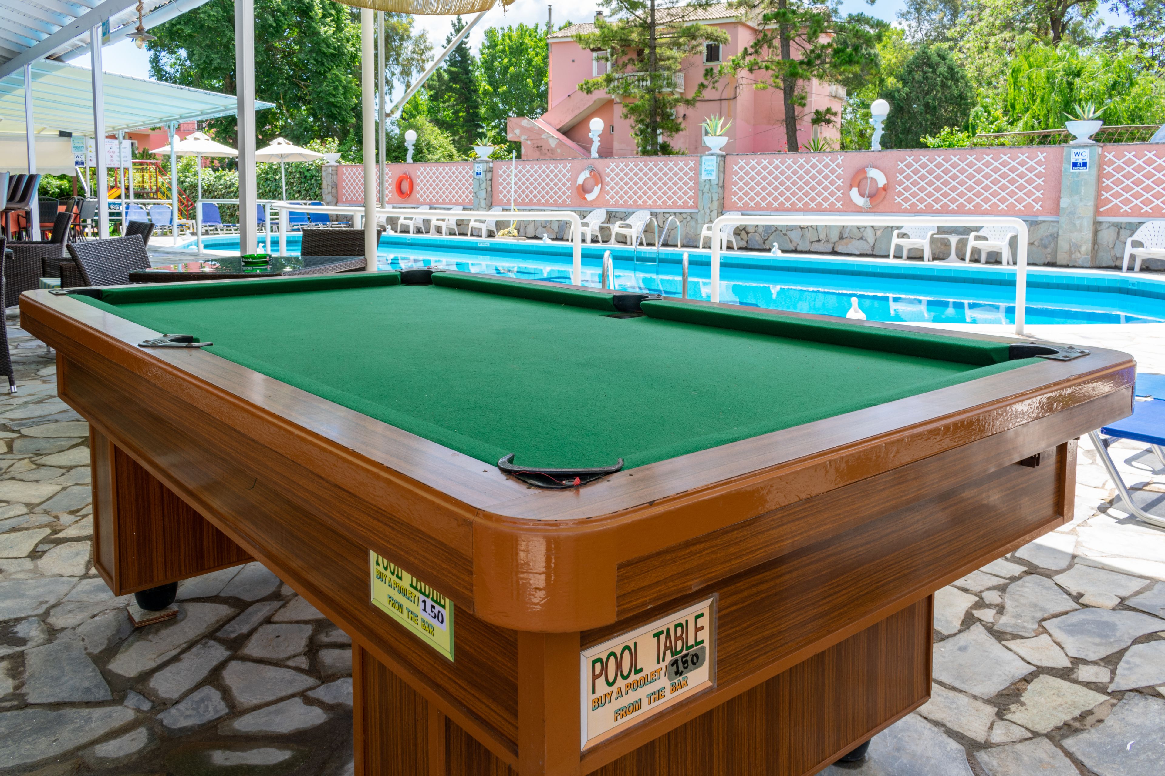 Una mesa de billar con superficie de fieltro verde, ubicada al aire libre, con una piscina y sillones de descanso al fondo.