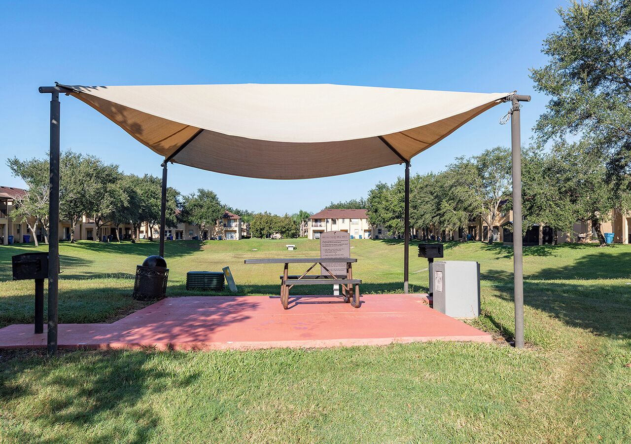 A covered outdoor seating area with a picnic table, surrounded by grass and trees.