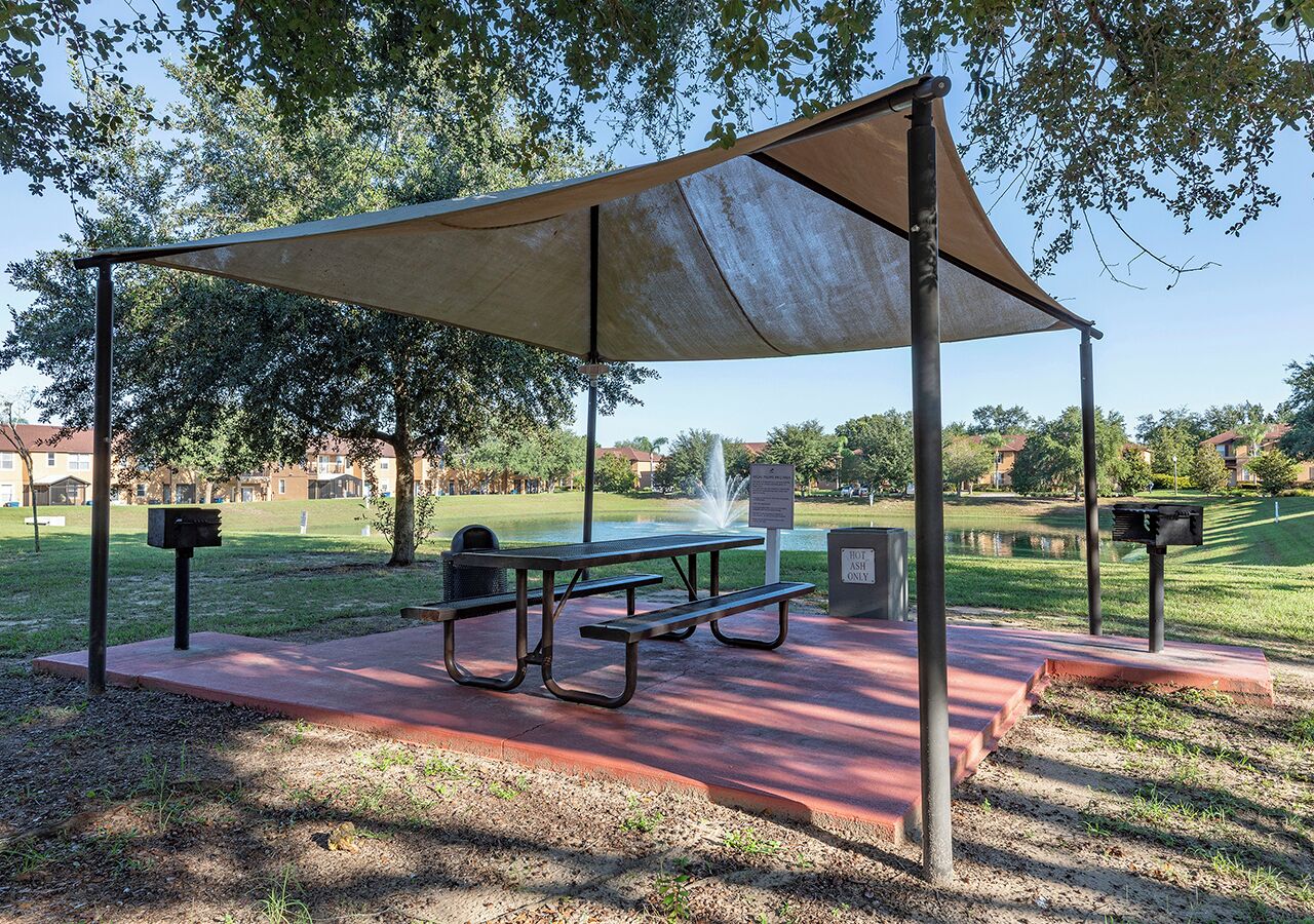 Uma área de piquenique com mesa e bancos sob um toldo sombreado, cercada por grama e árvores.