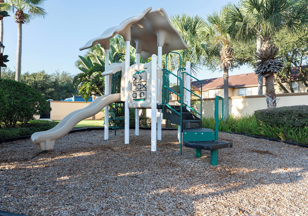 A playground with a slide and climbing structures, surrounded by gravel and palm trees.