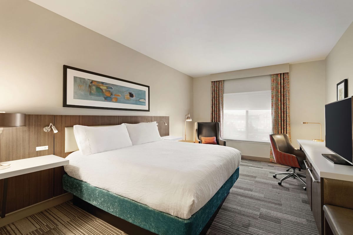 Habitación de hotel con una cama grande, un escritorio con computadora, una silla, un ventanal con cortinas y un cuadro en la pared.