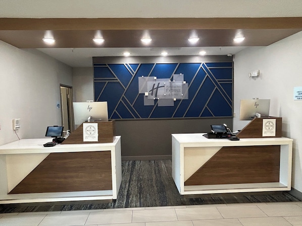 A reception area with two wooden reception desks, each with a computer monitor, phone, and a framed certificate. The wall behind features a geometric pattern and a sign for Holiday Inn Express & Suites. - Holiday Inn Express Hotel & Suites Grants - Milan by IHG (Grants)