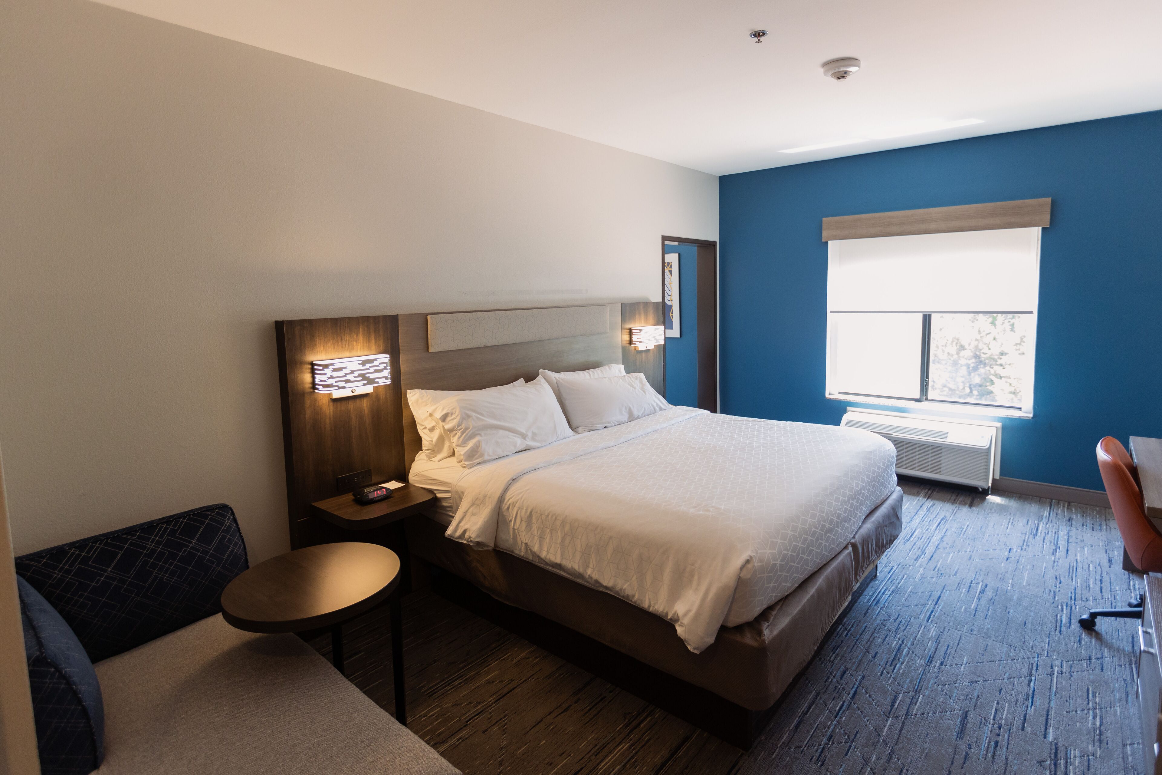 A hotel room with a large bed, a bedside table, a small round table, a sofa, and a window with blinds.