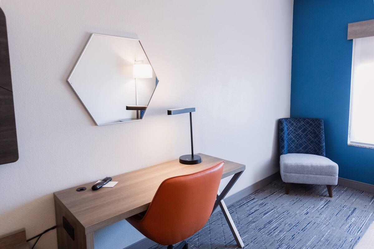 A hotel room with a wooden desk, a red chair, a small blue armchair, a mirror, and a lamp.