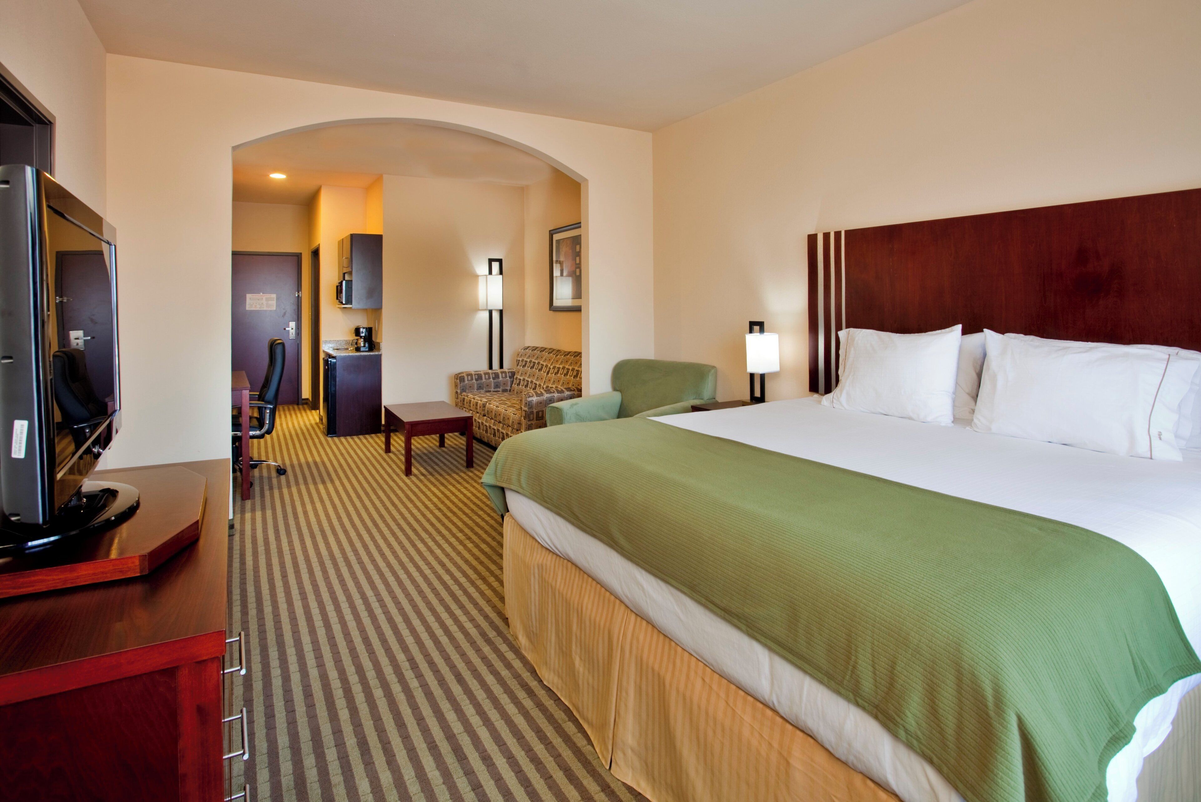 Habitación de hotel con una cama grande, un escritorio con ordenador, una silla, televisor y kitchenette.