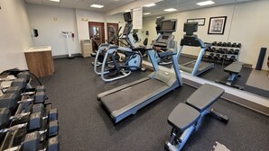Une salle de sport équipée de tapis de course, de vélos elliptiques et d’un banc. On y trouve également des haltères et un miroir fixé au mur.
