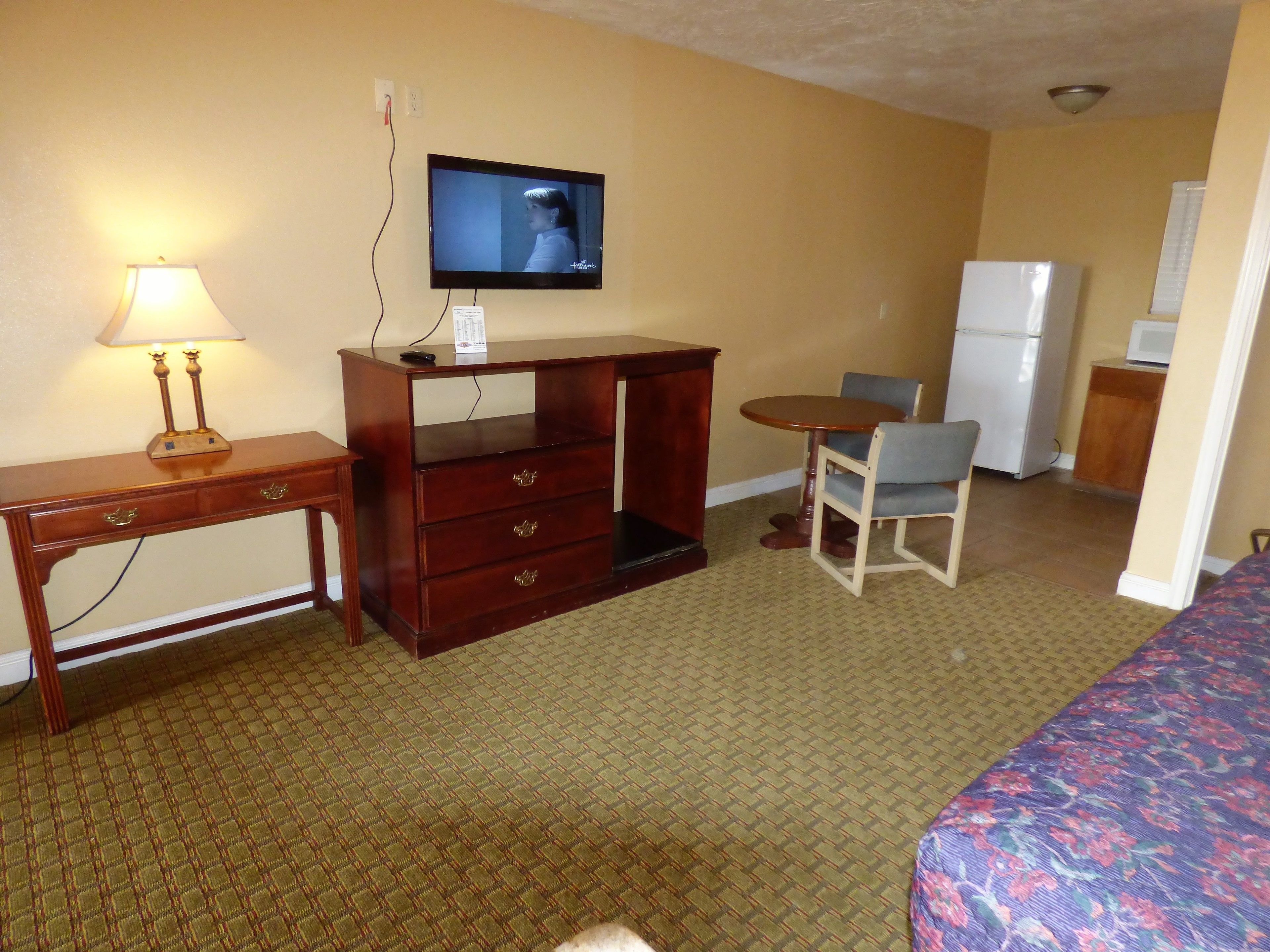 Een hotelkamer met een bed, een bureau met lamp, een televisie, een kleintje tafeltje met stoel, en een koelkast.