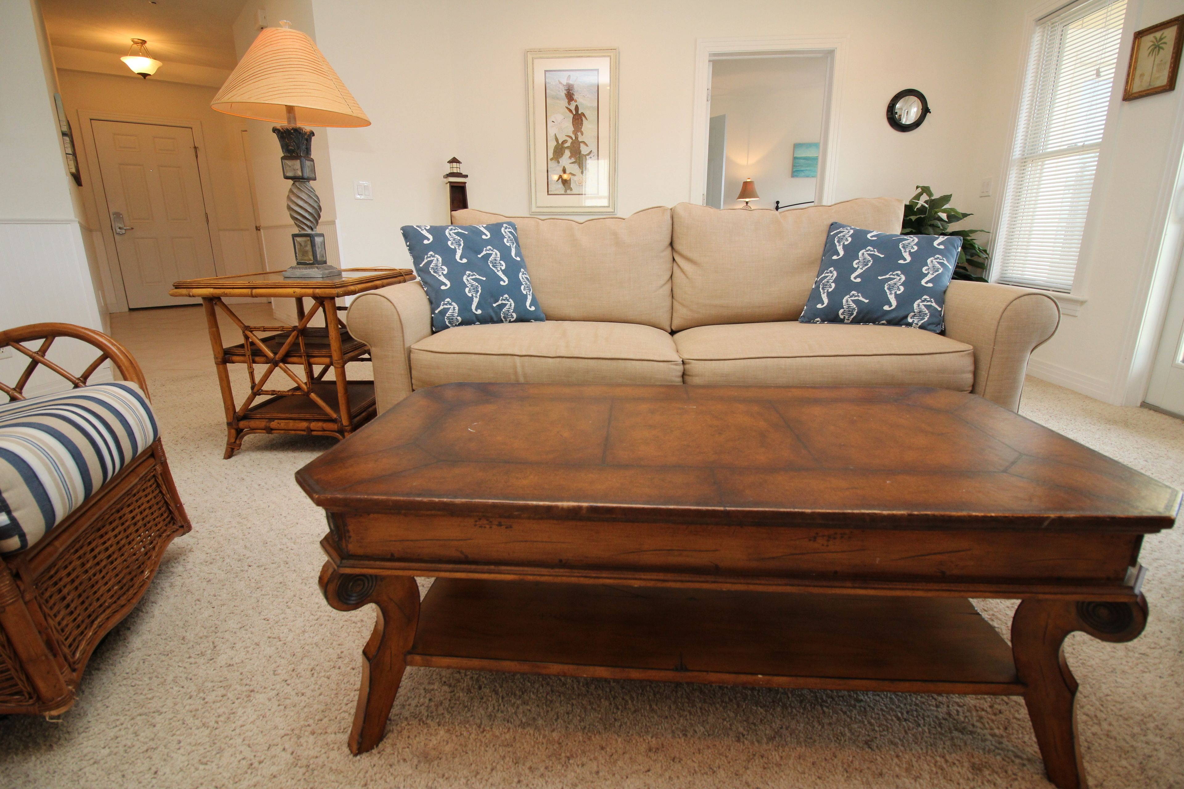 A living room with a wooden coffee table, a beige sofa with patterned cushions, a wicker chair, and a small side table with a lamp.