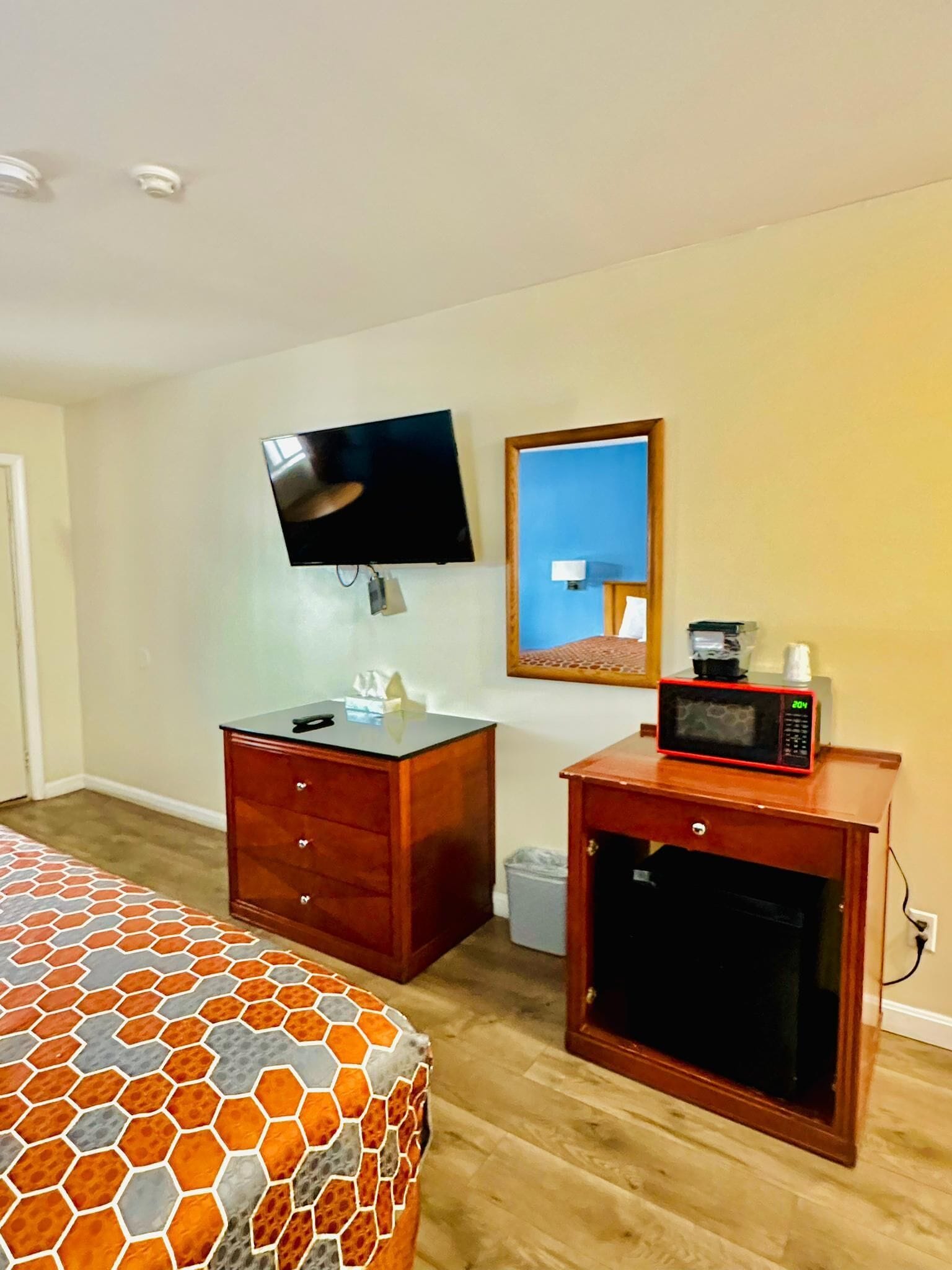 Una habitación de hotel con un aparador de madera, un televisor montado en la pared, un microondas sobre una mesita y una colcha estampada.