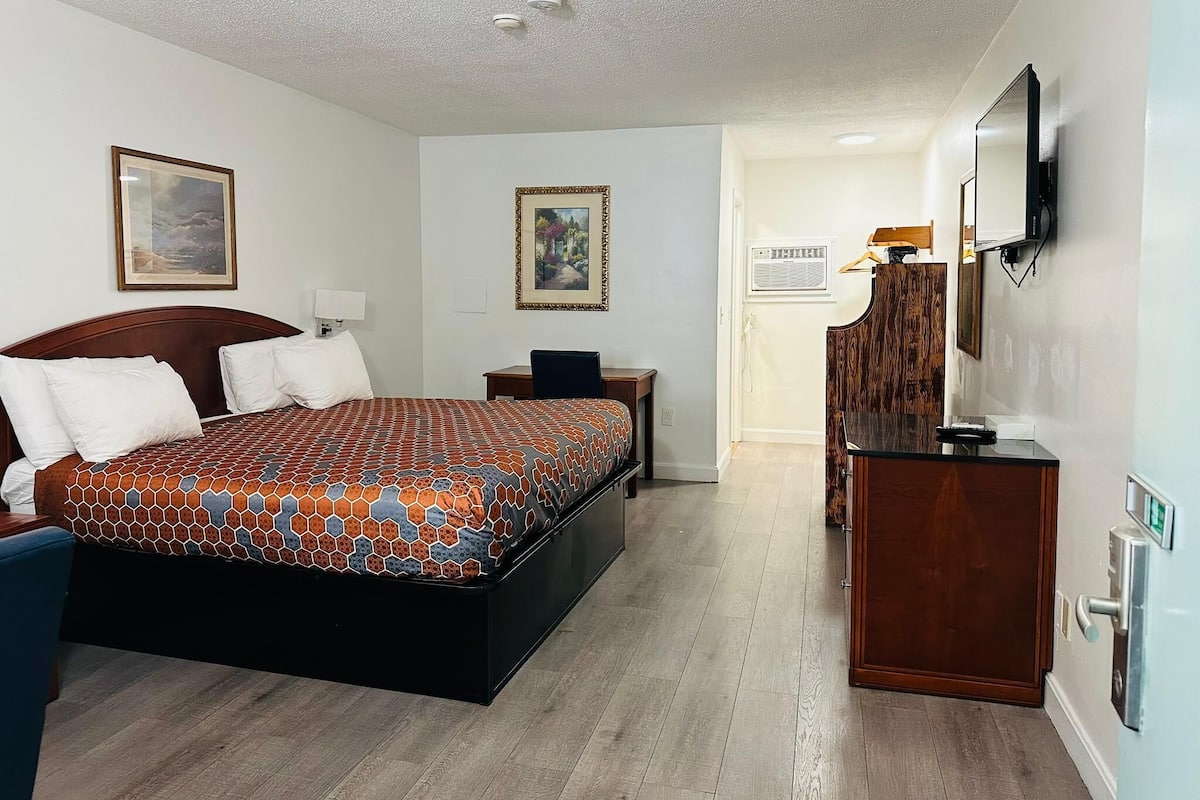 Ein Hotelzimmer mit einem großen Bett, einem Schreibtisch, einem Sessel, einer Kommode, einem Fernseher und einer Tür zu einem weiteren Raum.
