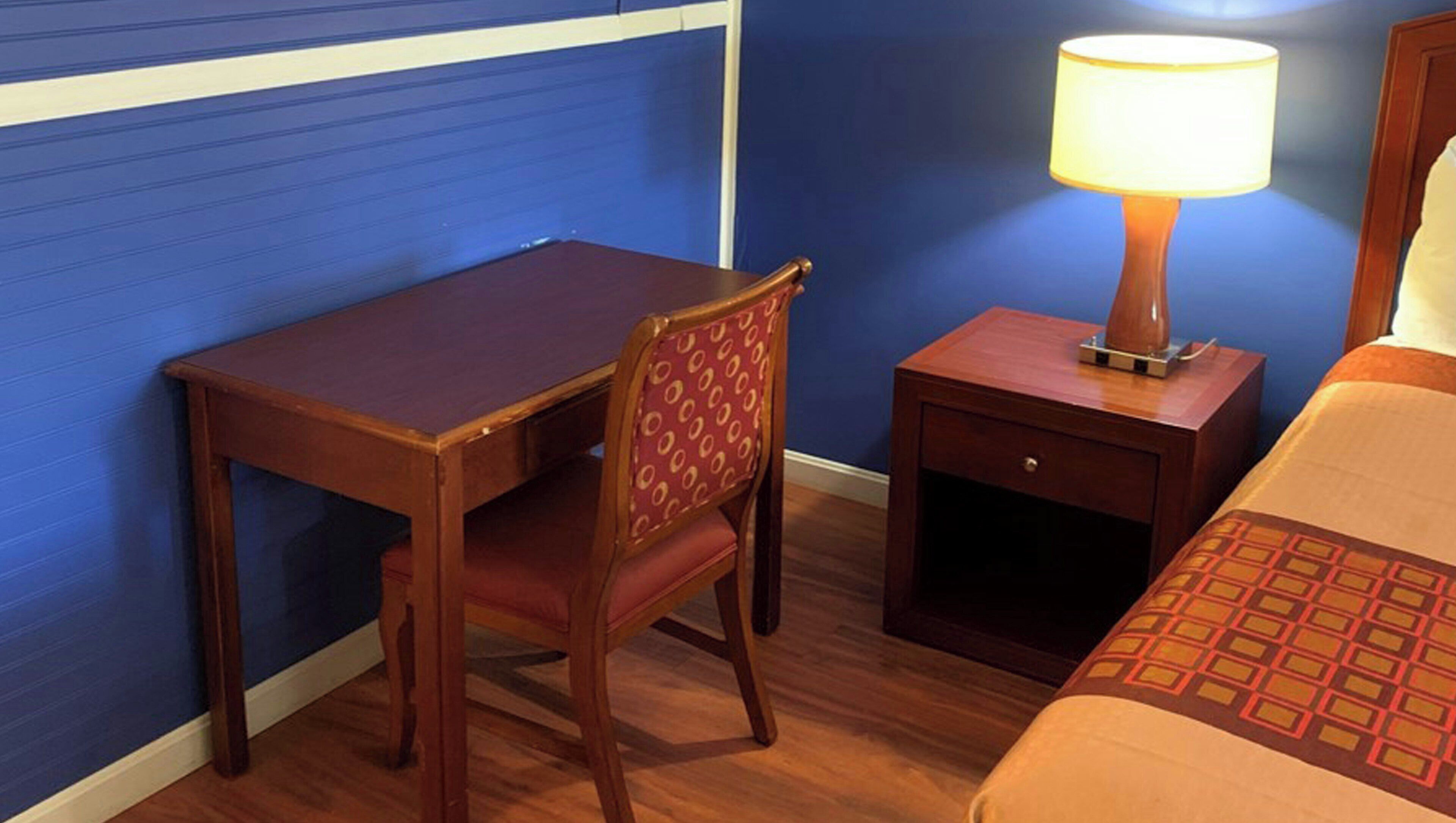 Habitación de hotel con escritorio de madera, una silla, mesita de noche con lámpara y cama con colcha estampada.