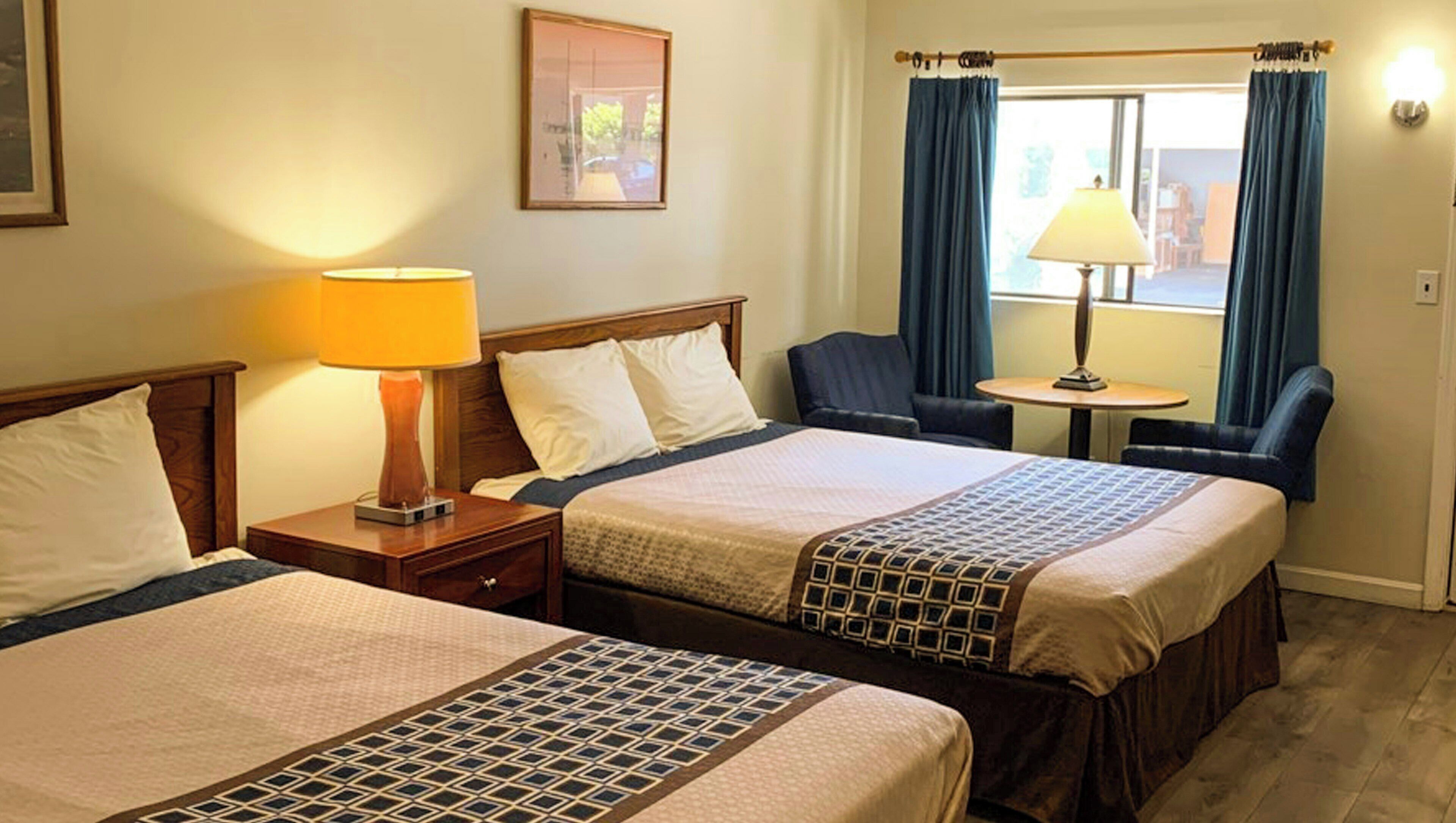 A hotel room with two beds, a nightstand with a lamp, a chair, a small table, and a window with curtains.