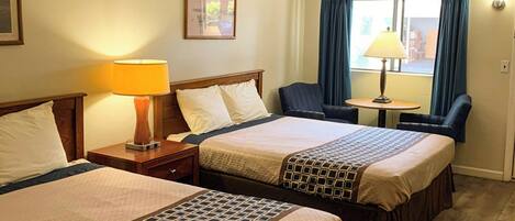 A hotel room with two beds, a nightstand with a lamp, a chair, a small table, and a window with curtains.