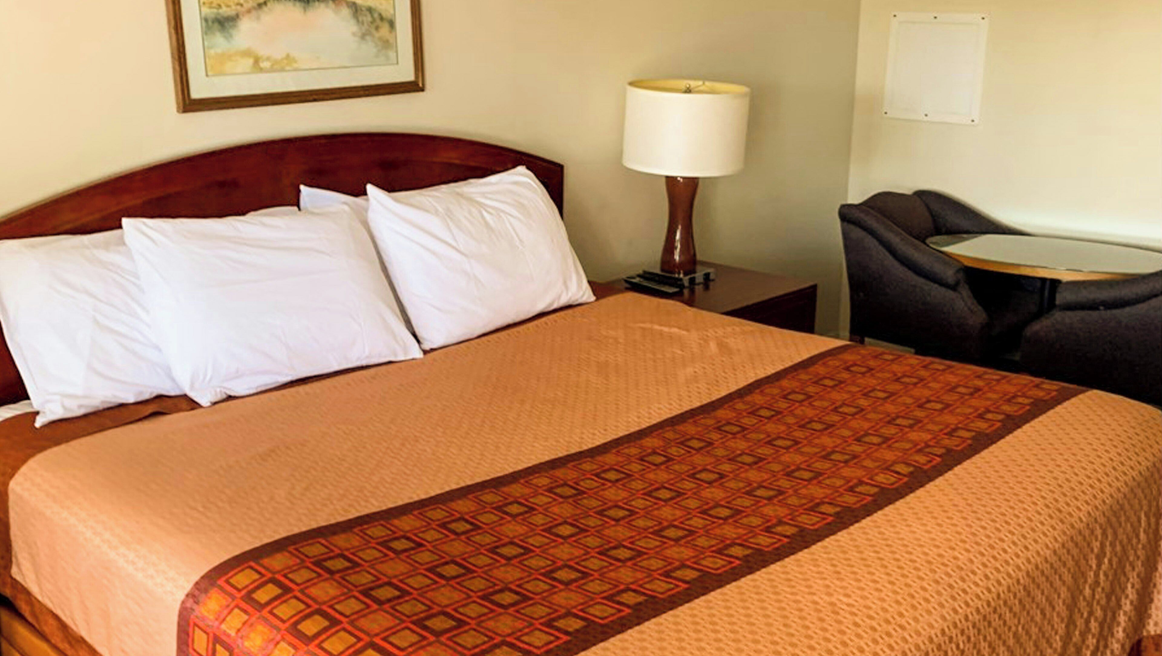 A hotel room with a bed, a nightstand with a lamp, a small table, and a chair.
