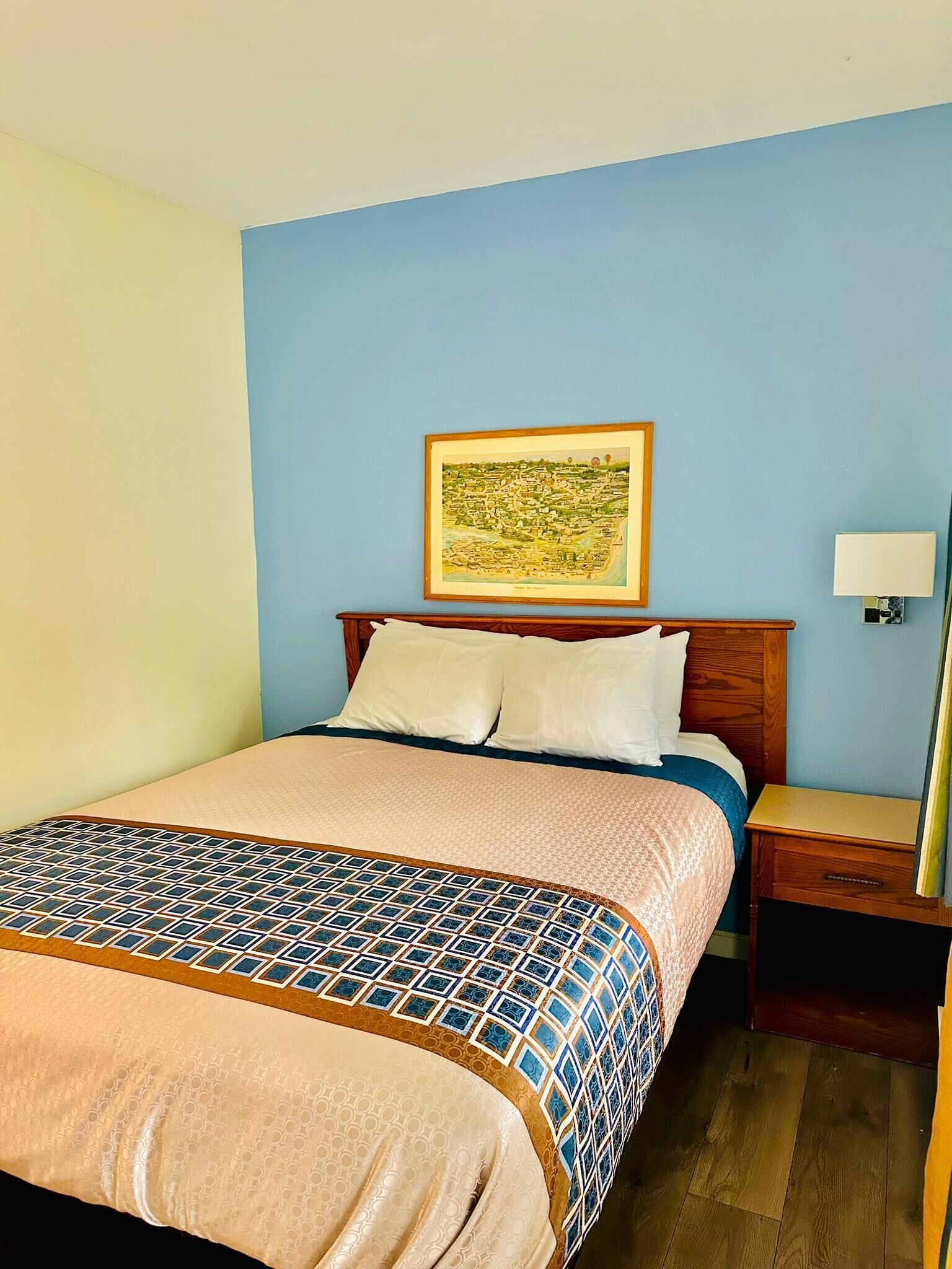 Una habitación de hotel con una cama, una mesita de noche, una lámpara y un cuadro enmarcado con una vista urbana.