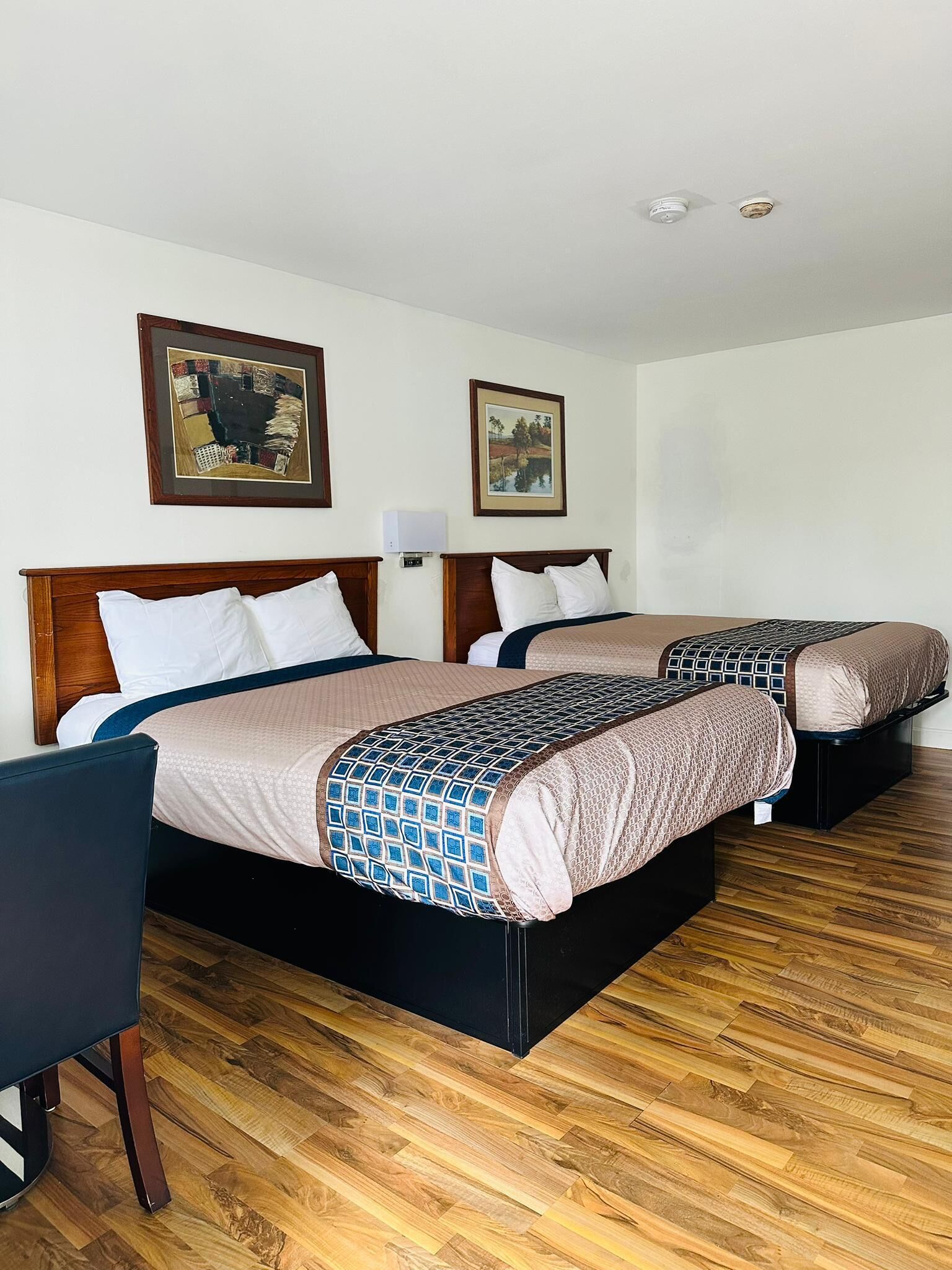 A hotel room with two beds, wooden headboards, a wooden floor, and two framed pictures on the wall.