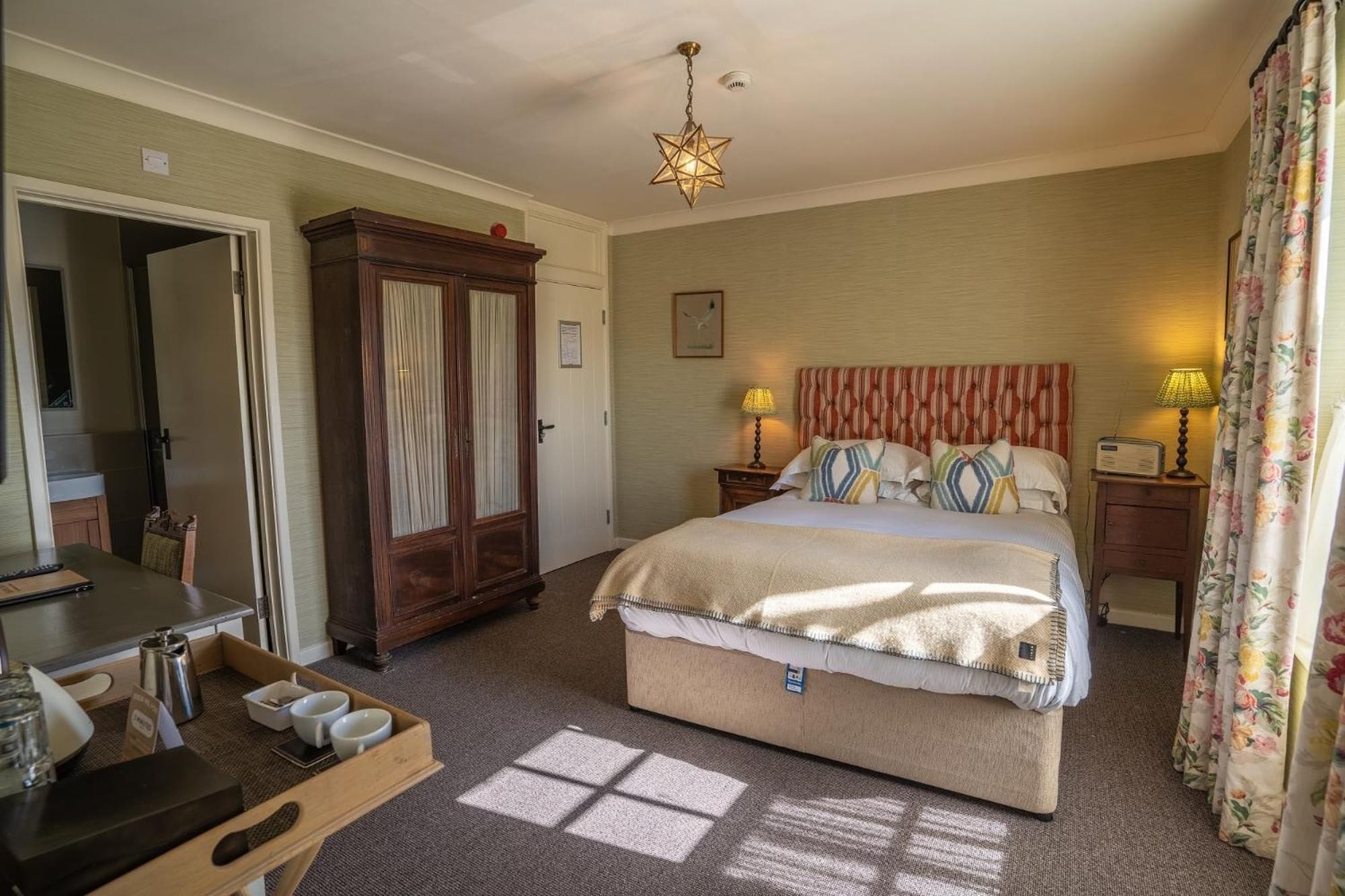 A bedroom with a bed, bedside lamps, a wooden wardrobe, a desk with a chair, and a window with curtains.