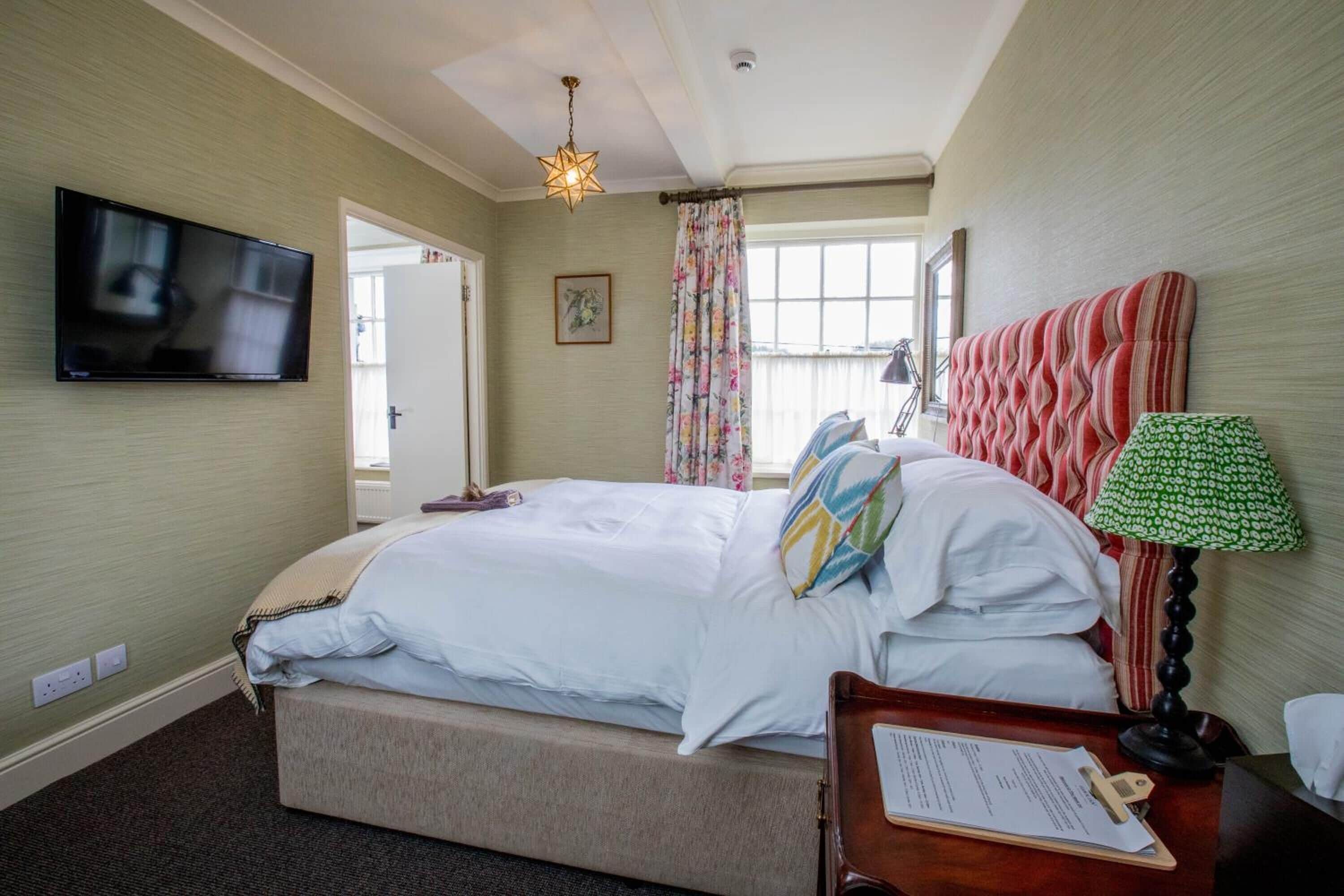 A hotel room with a bed, a flat-screen TV, a desk with a lamp, and a window with curtains.