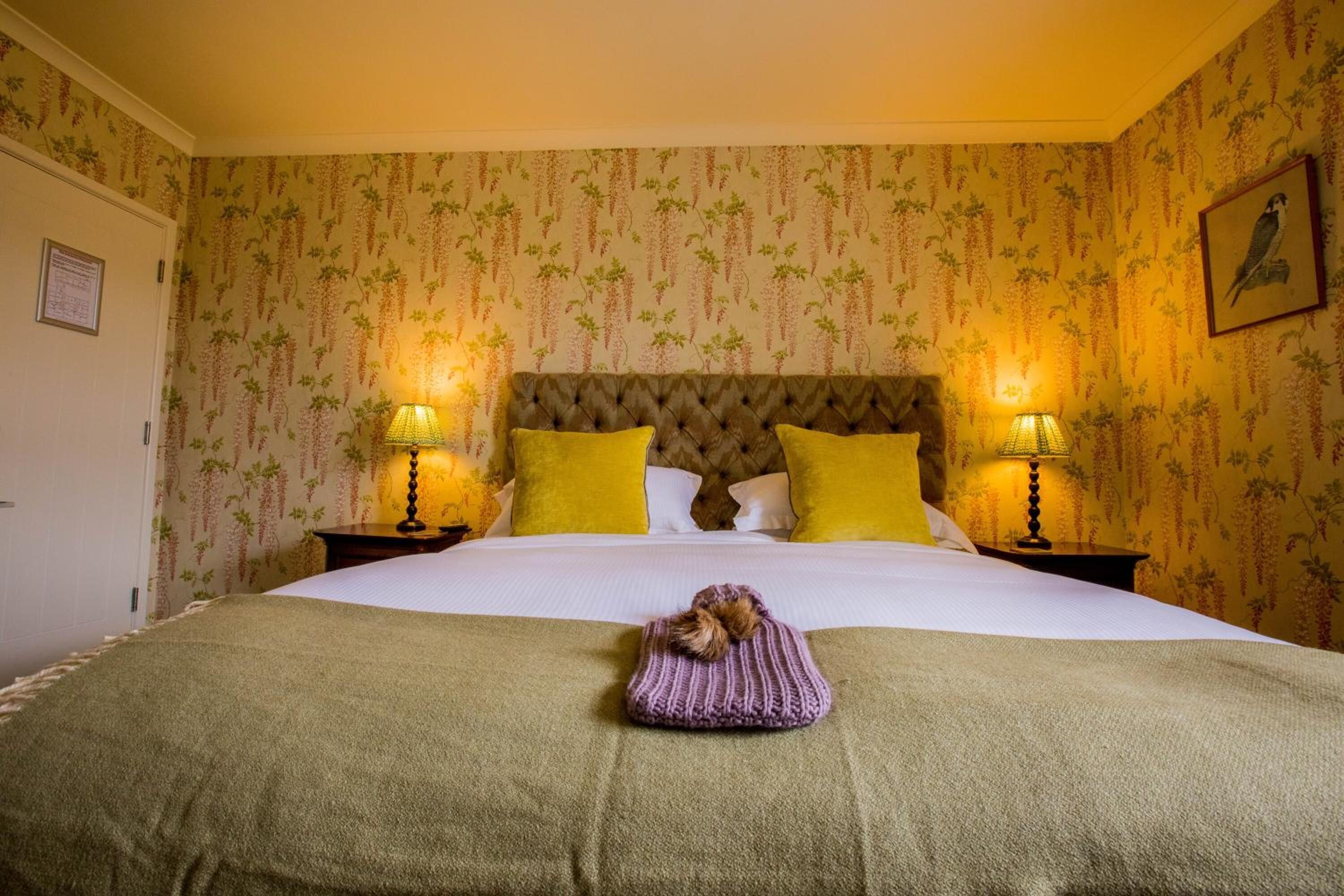 A neatly made bed with a green blanket, two yellow pillows, and a tufted headboard. There are two bedside lamps on either side of the bed.