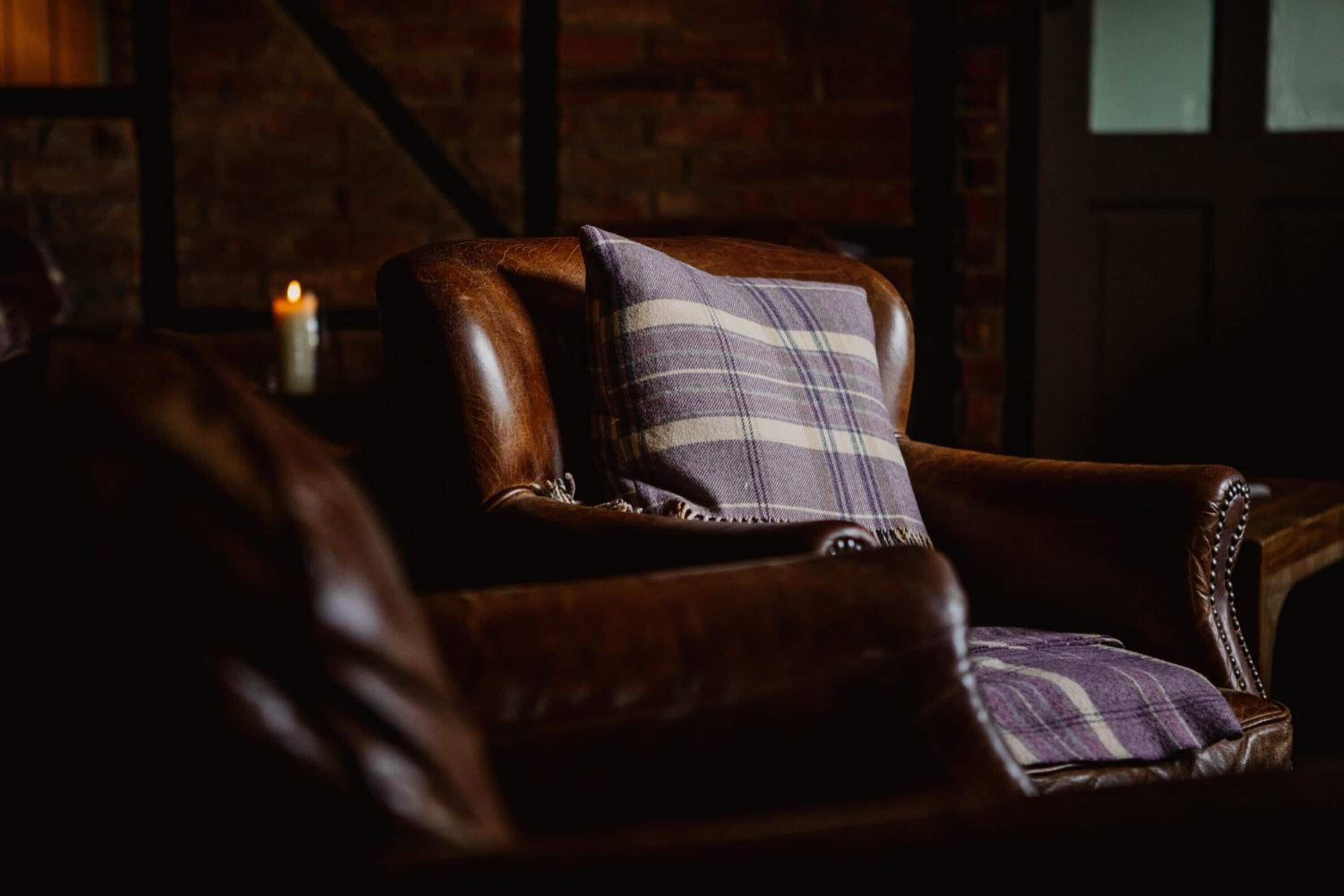 A leather armchair with a plaid cushion in a dimly lit room with a brick wall and a lit candle.