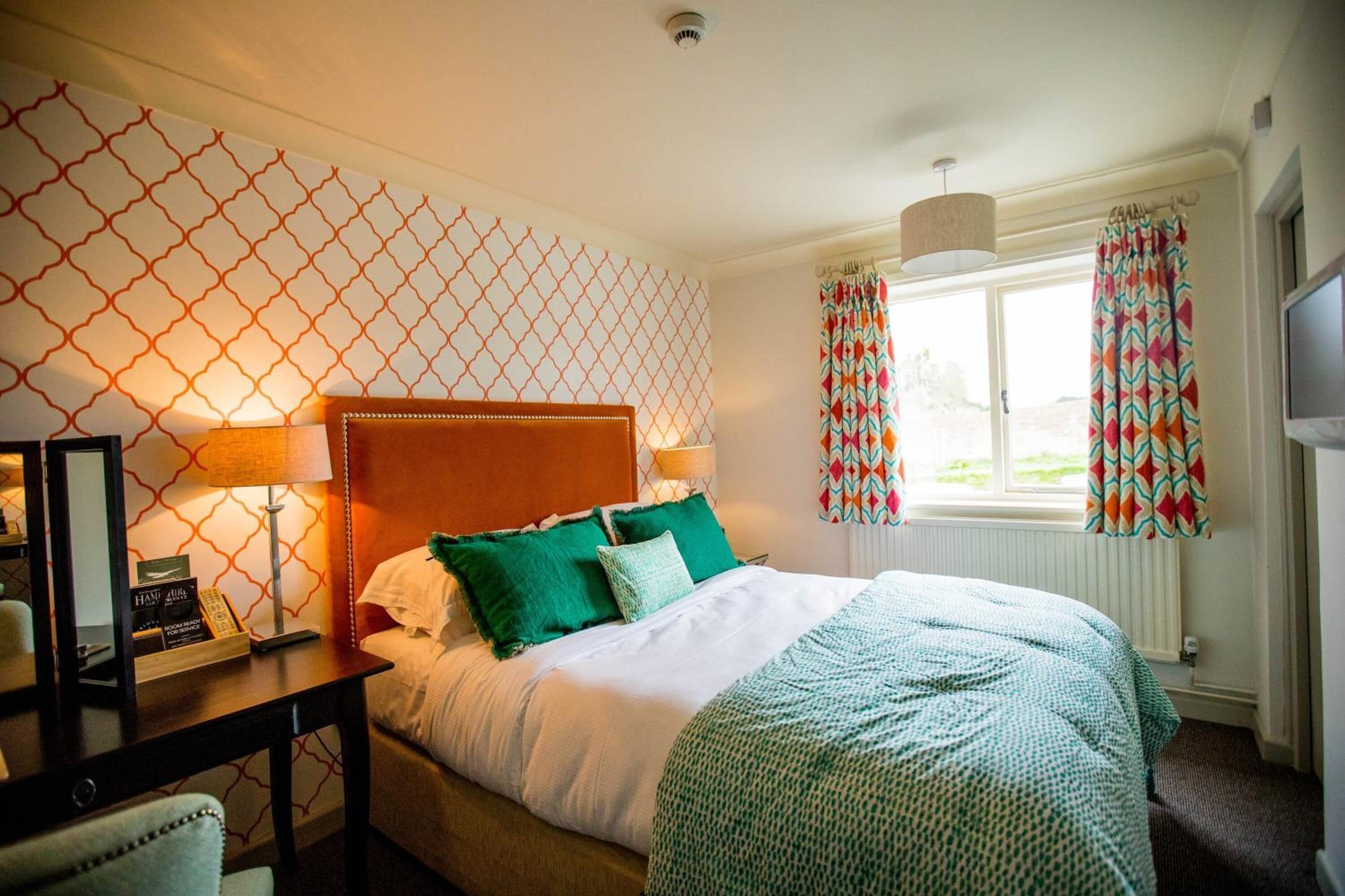 A hotel room with a bed, a desk with a lamp, a chair, a window with curtains, and a wall with a geometric pattern.