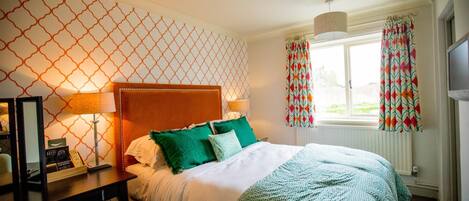 A hotel room with a bed, a desk with a lamp, a chair, a window with curtains, and a wall with a geometric pattern.