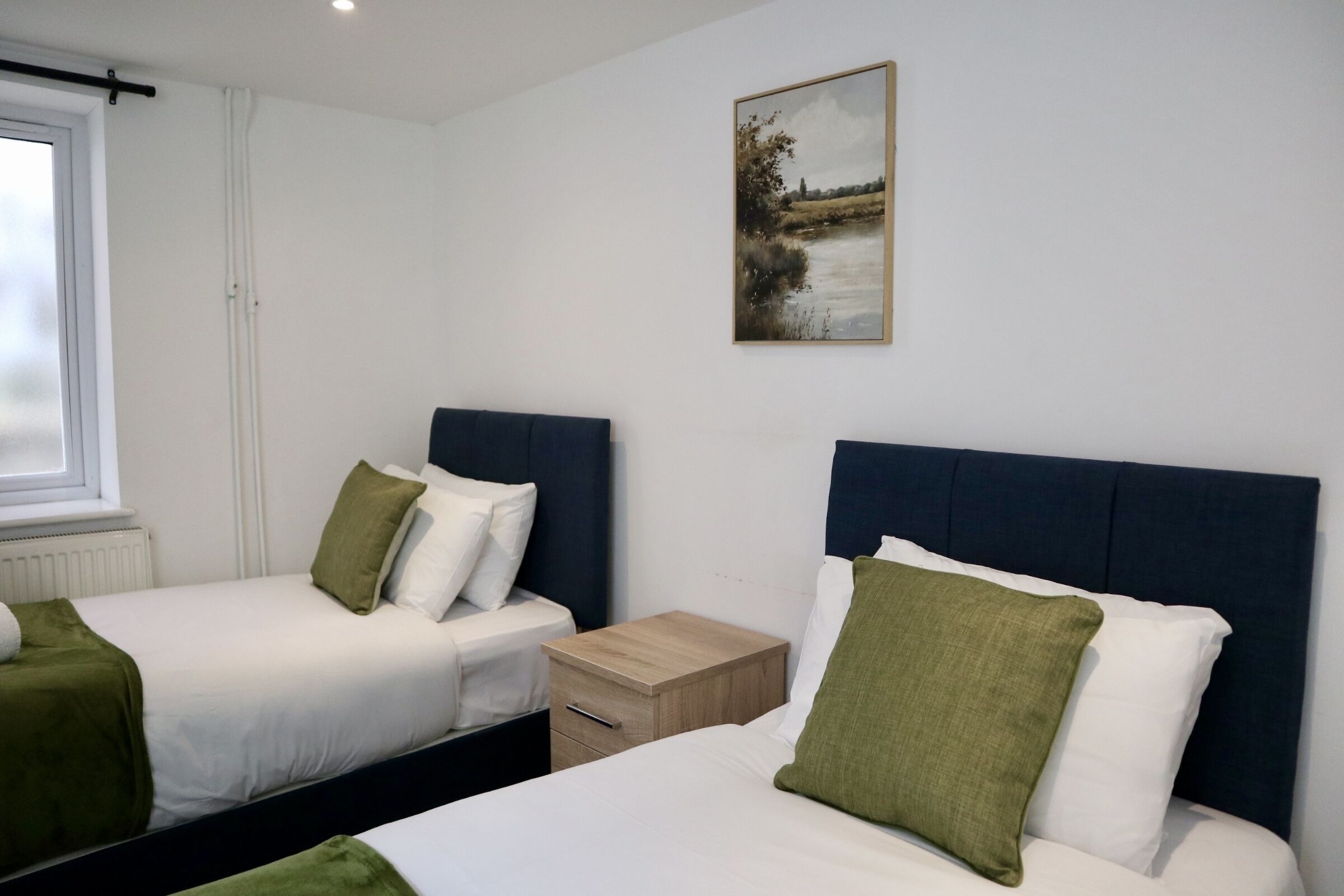 A bedroom with two beds, a nightstand, a window, and a painting on the wall.