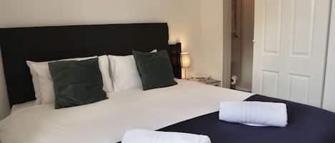 A neatly made bed with white and dark green pillows, a dark blue bedspread, and two rolled white towels.