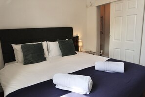 A neatly made bed with white and dark green pillows, a dark blue bedspread, and two rolled white towels. - City Centre Guest House - Gloucester Park (Gloucester)