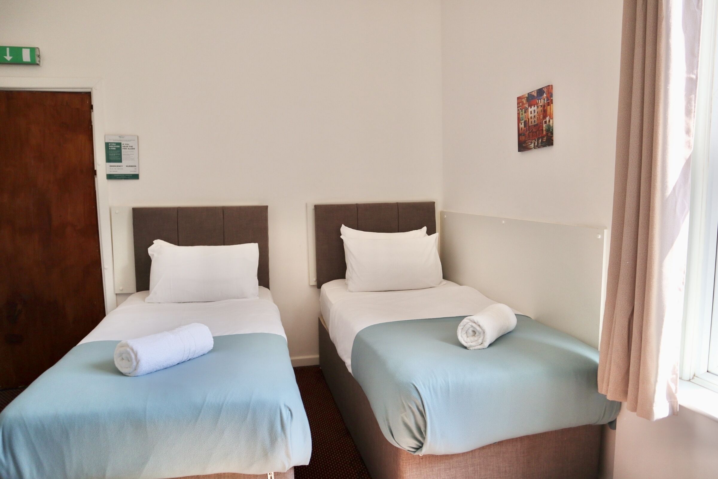 Habitación de hotel con dos camas individuales, una puerta de madera, un letrero de salida verde y un pequeño cuadro en la pared.