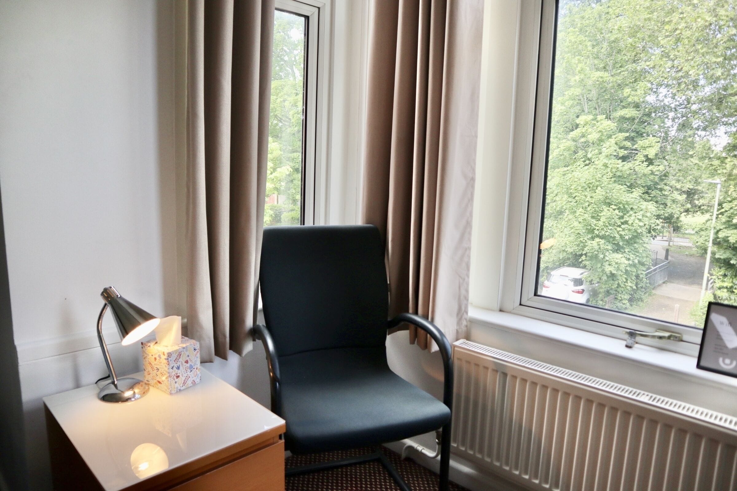 Una habitación con una silla cerca de una ventana, una mesita de noche con una lámpara y una caja de pañuelos.
