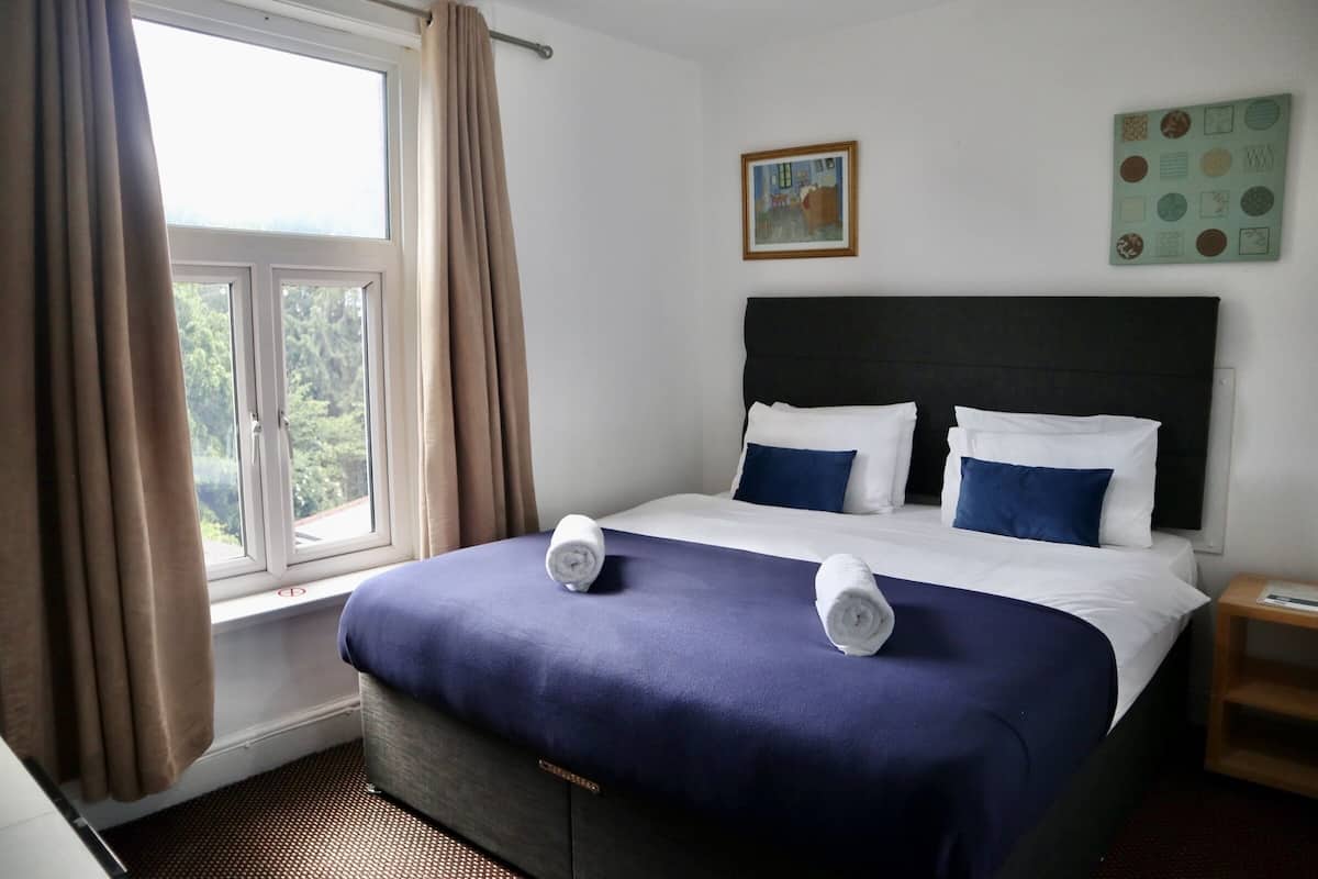 A hotel room with a bed, a window with curtains, a painting on the wall, and a small wooden side table.