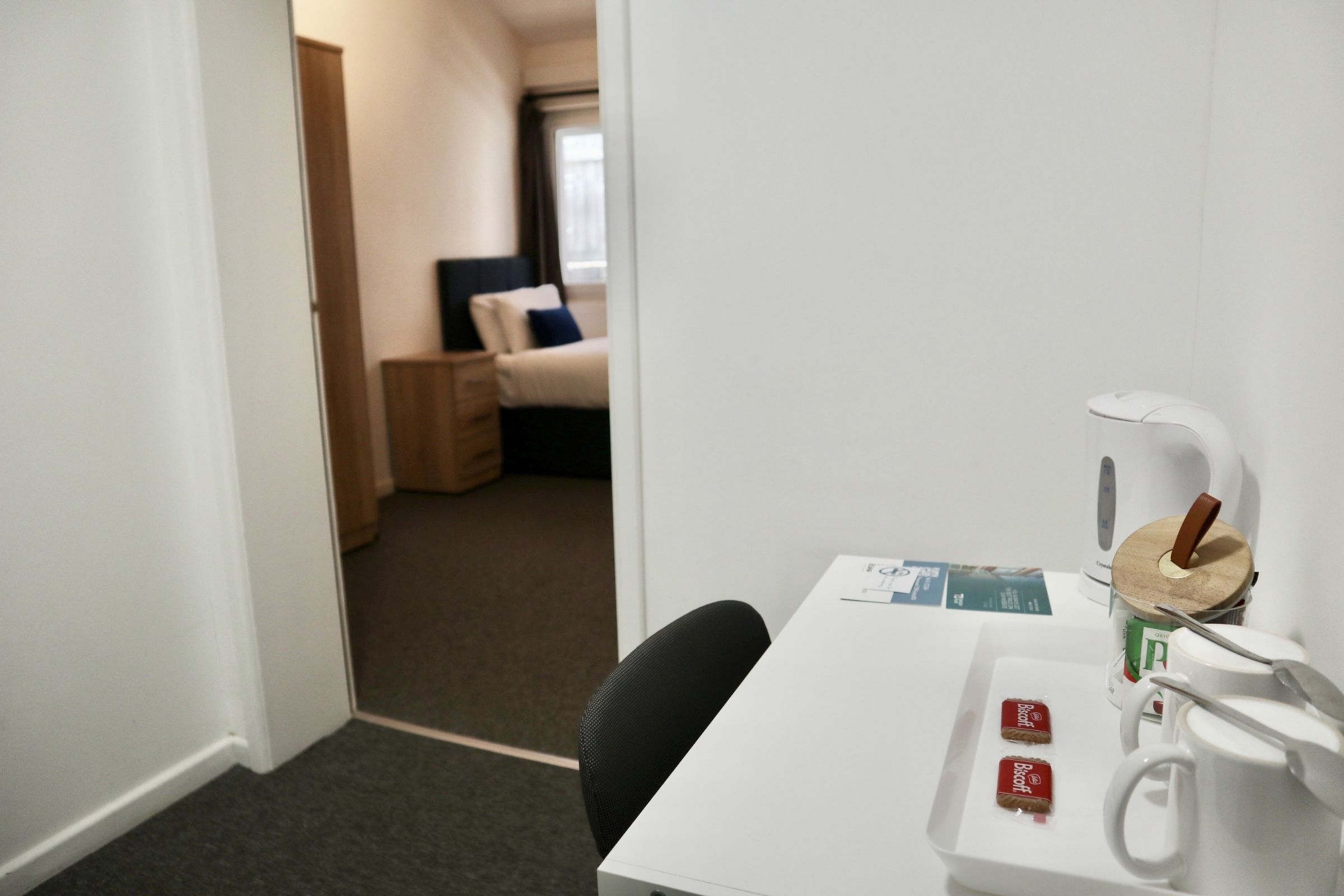 Habitación de hotel con un escritorio blanco, una silla, una cama con almohada azul y una mesita de noche con lámpara.