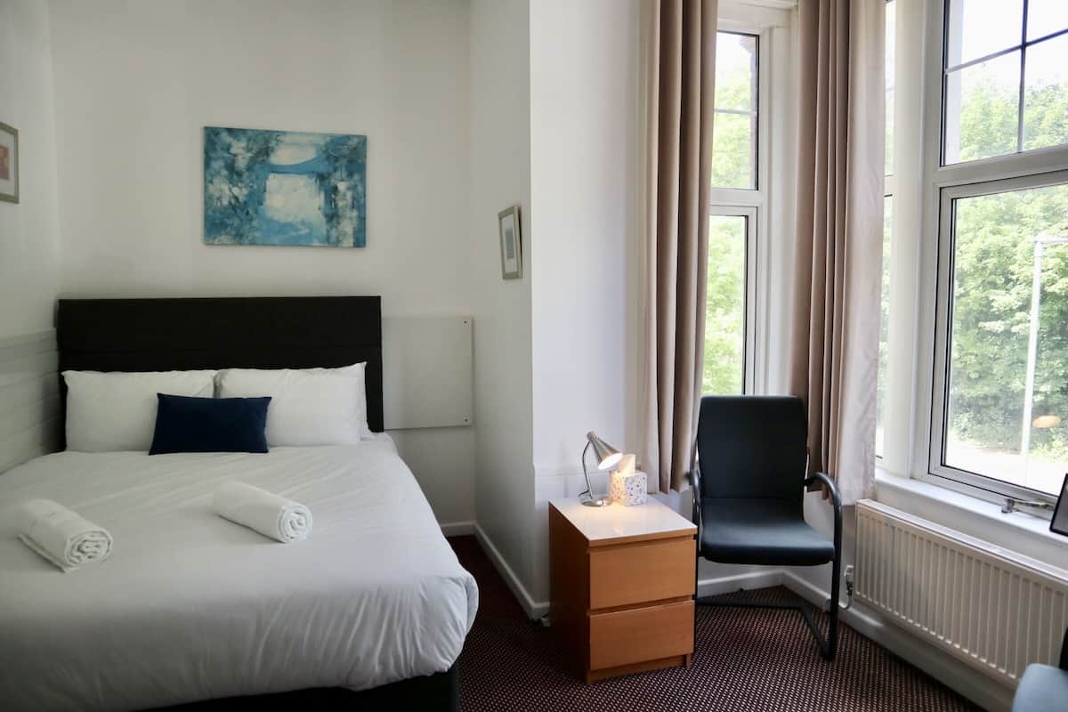 A hotel room with a bed, a nightstand, a chair, a window, and a painting on the wall.