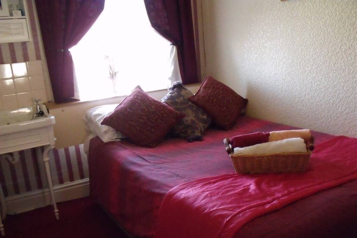 A bedroom with a bed, pillows, a nightstand, a window with curtains, and a basket on the bed.