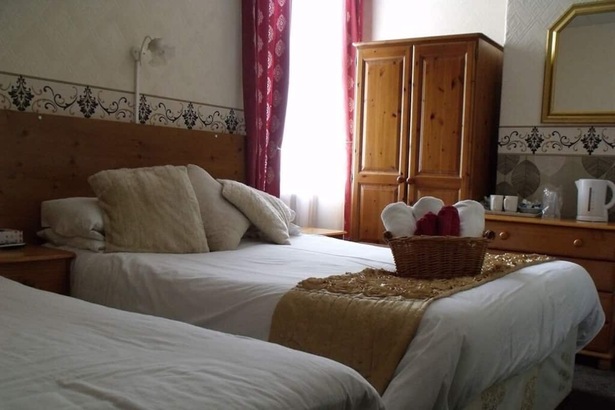 A hotel room with two beds, a wooden wardrobe, a mirror, and a window with red curtains.