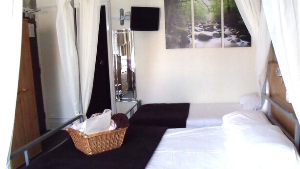 A bedroom with two beds, a mirror, a television, and a painting of a forest scene. - The Derby Hotel (Blackpool)