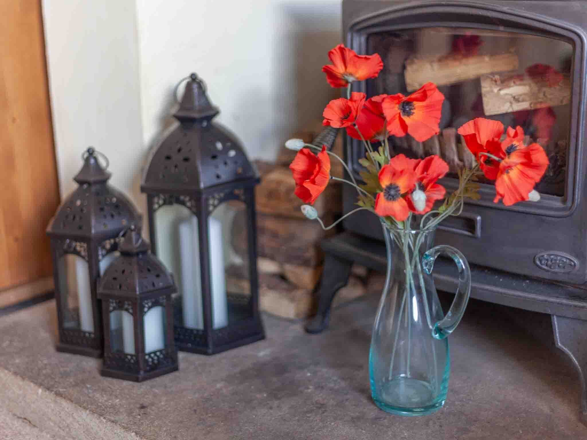 A glass vase with red flowers on a stone floor, next to two black lanterns and a wood-burning stove.