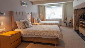 Une chambre avec deux lits, une table de chevet en bois, une chaise, une cheminée et une fenêtre avec des stores.