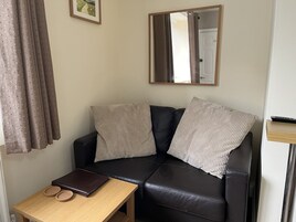 Un pequeño salón con un sofá de cuero negro, dos almohadas color beige, una mesita de madera con una laptop y cuencos, un espejo en la pared y una cortina estampada con lunares.