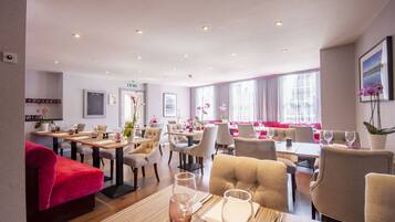 A spacious dining area with multiple tables and chairs, a red sofa, and large windows providing natural light.