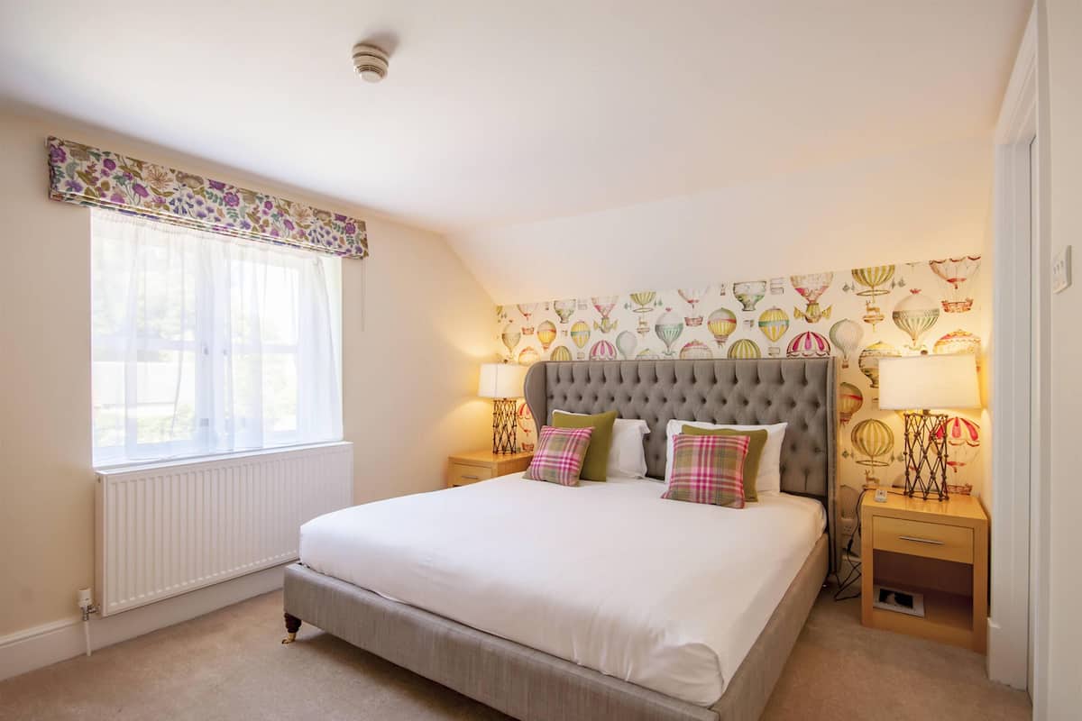 A bedroom with a large bed, two bedside tables with lamps, a radiator, and a window with sheer curtains.
