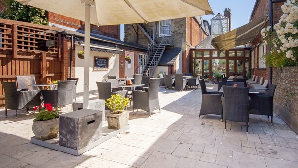 An outdoor patio with wicker furniture, a large umbrella, and a brick building in the background. - The Highworth Hotel (Swindon)