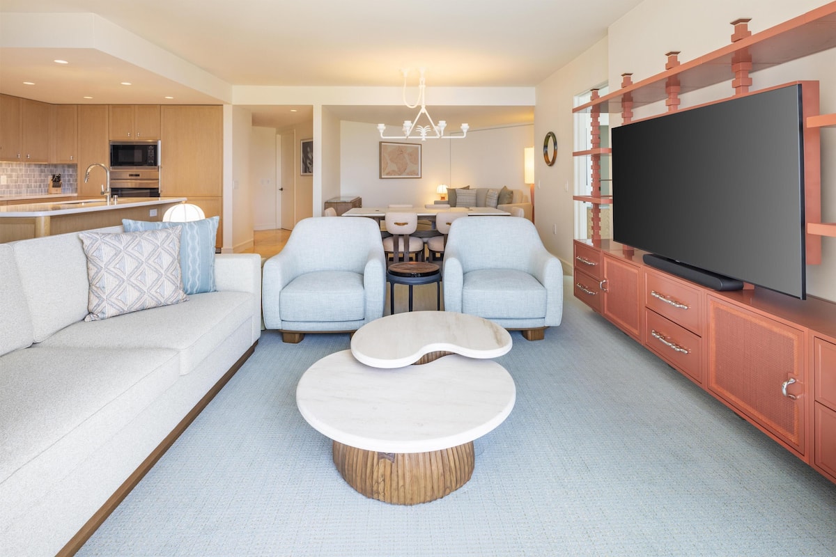 Een moderne woonkamer met een bank, twee fauteuils, een salontafel, een televisie en een keuken op de achtergrond.