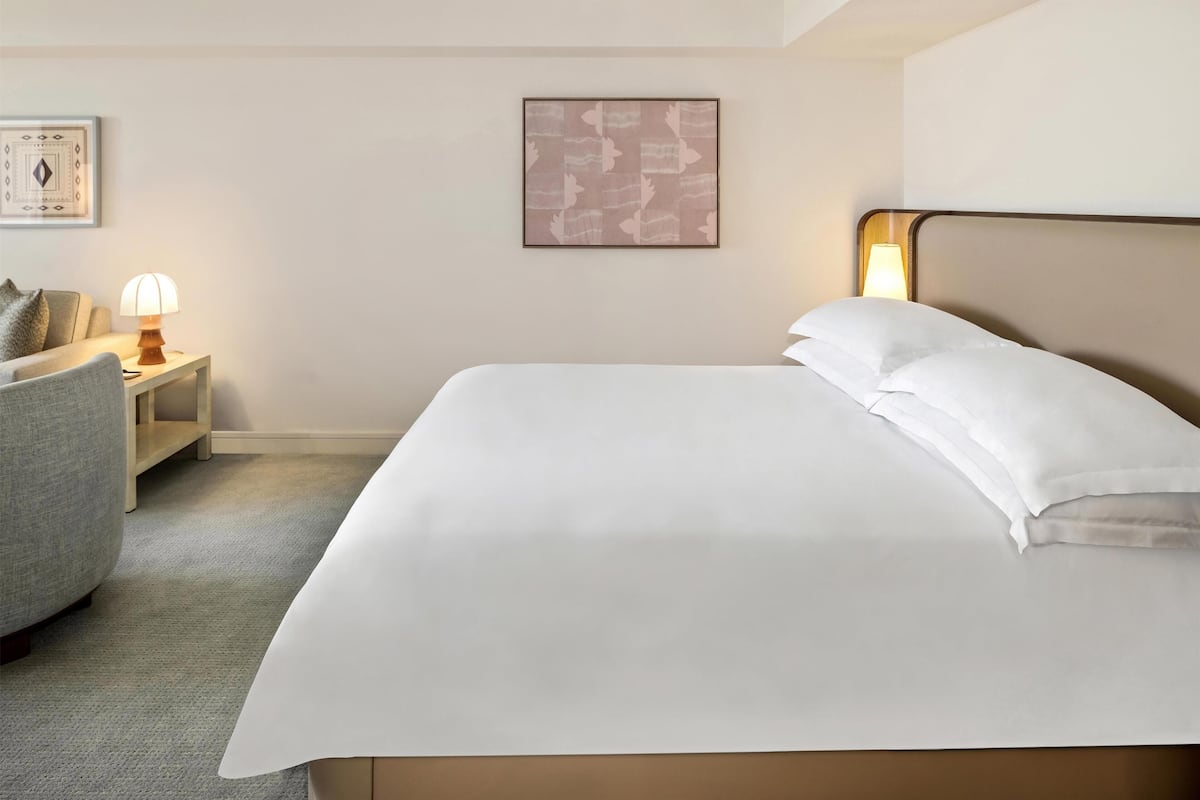 Een net opgemaakt bed met witte lakens, een bedlampje, een stoel en een kunstwerk in een lijst aan de muur.