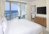A hotel room with a large bed, a TV, a wooden dresser, and a balcony with a view of the ocean.