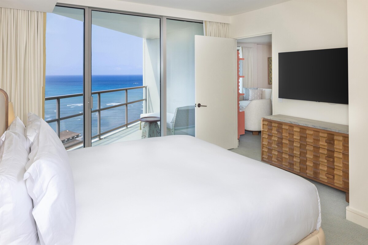 A hotel room with a large bed, a TV, a wooden dresser, and a balcony with a view of the ocean.