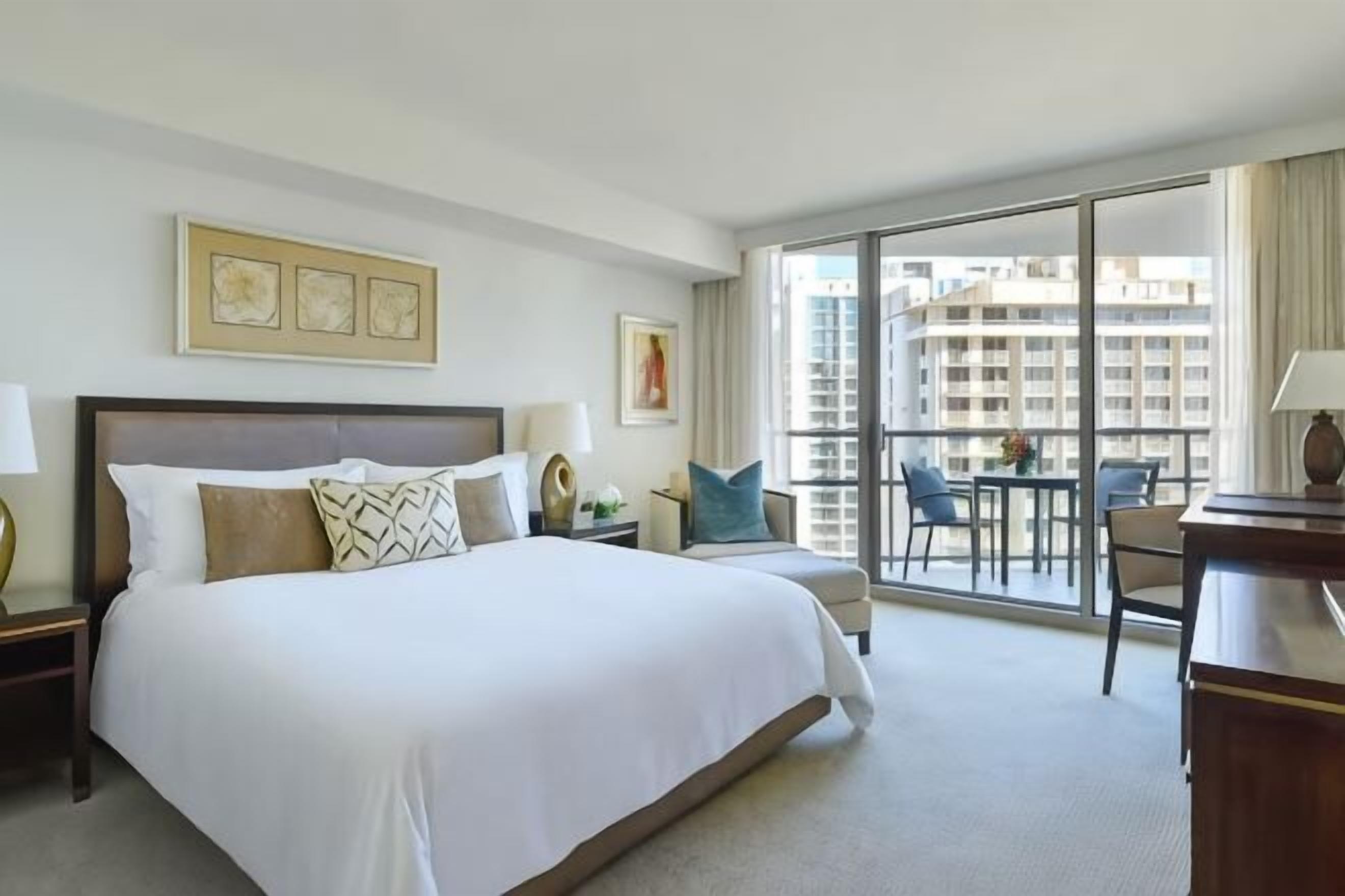 Habitación de hotel con una cama grande, un escritorio, una silla y vistas de la ciudad a través de amplias ventanas.