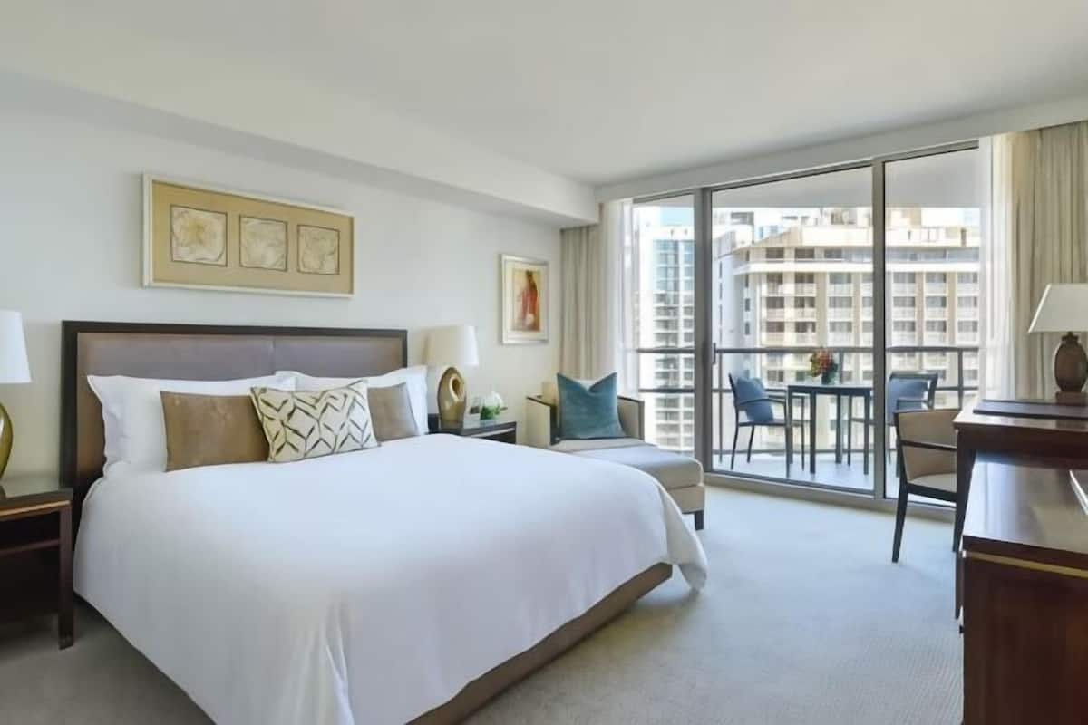 A hotel room with a large bed, a desk, a chair, and a view of the city through large windows.